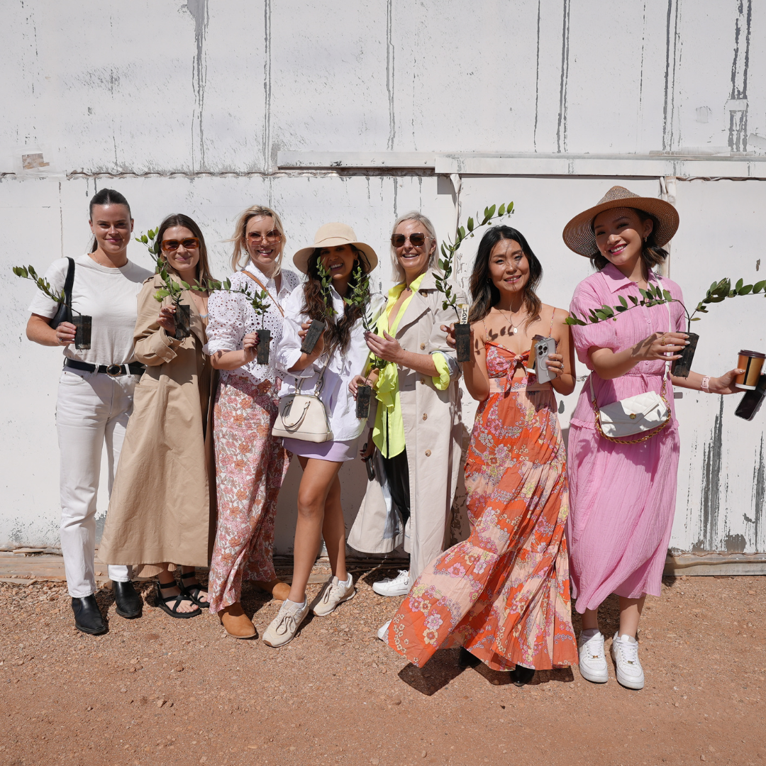 Group of people in various outfits standing against a white wall holding green branches.