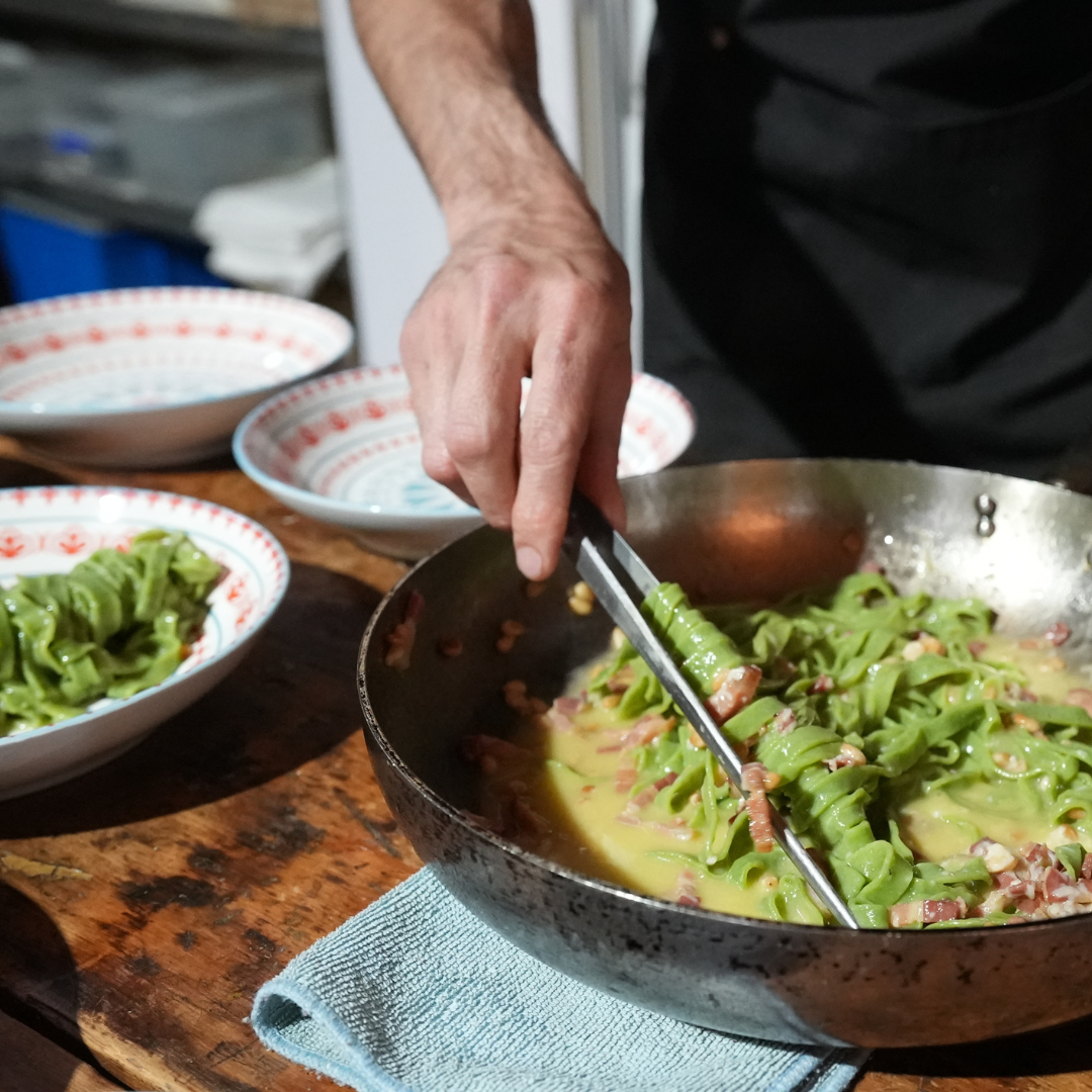 Person serving green pasta with tongs from a skillet, with plated dishes in the background.