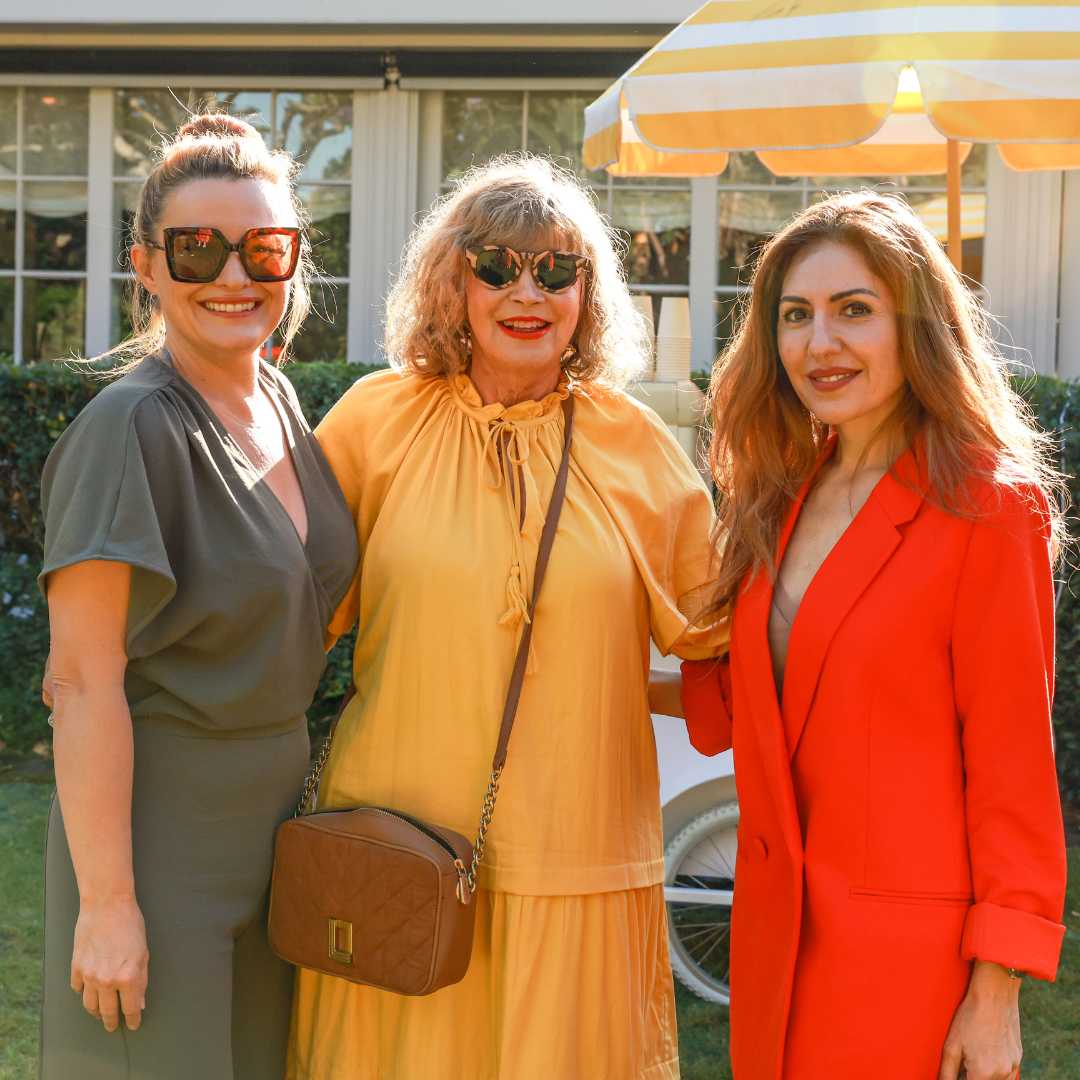 Three women posing together outdoors, two in vibrant dresses, one in casual attire.