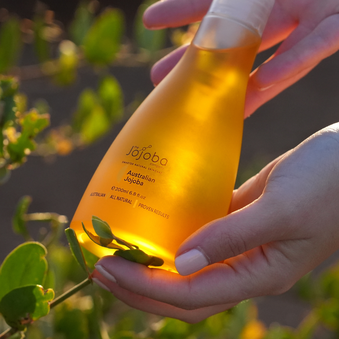 Hand holding a bottle of Australian Jojoba oil with foliage in the background, illuminated by soft sunlight.