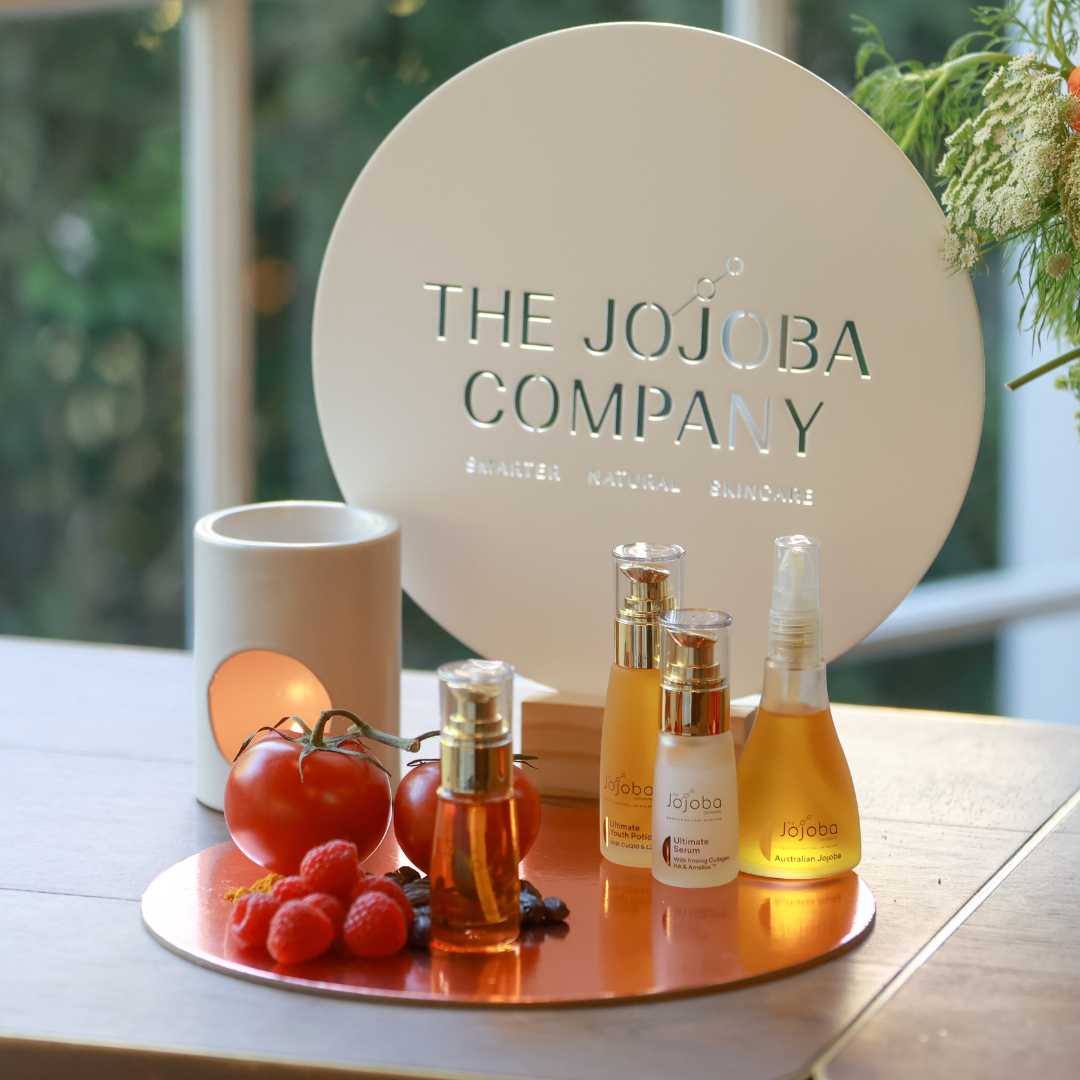 Cosmetic products from The Jojoba Company on a copper tray with tomatoes and berries, next to a candle.