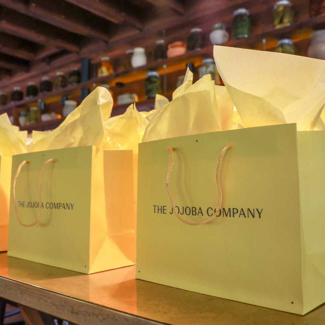 Green shopping bags with "THE JOJOBA COMPANY" logo on a counter, with shelves in the background.