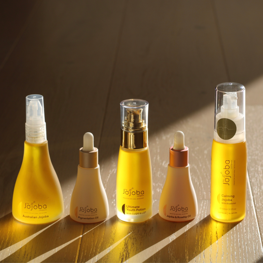 Five Jojoba skincare bottles on a wooden surface with sunlight casting shadows.