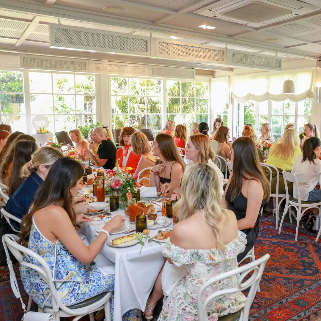 A group of people dining in a bright room with large windows and floral arrangements.