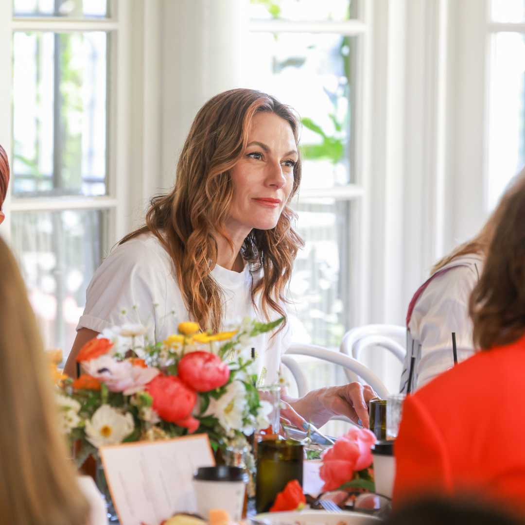 A person in a white shirt seated at a table with vibrant flowers, conversing with others.