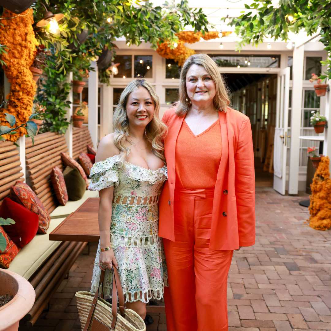 Two people in stylish outfits standing in a garden patio surrounded by plants and flowers.