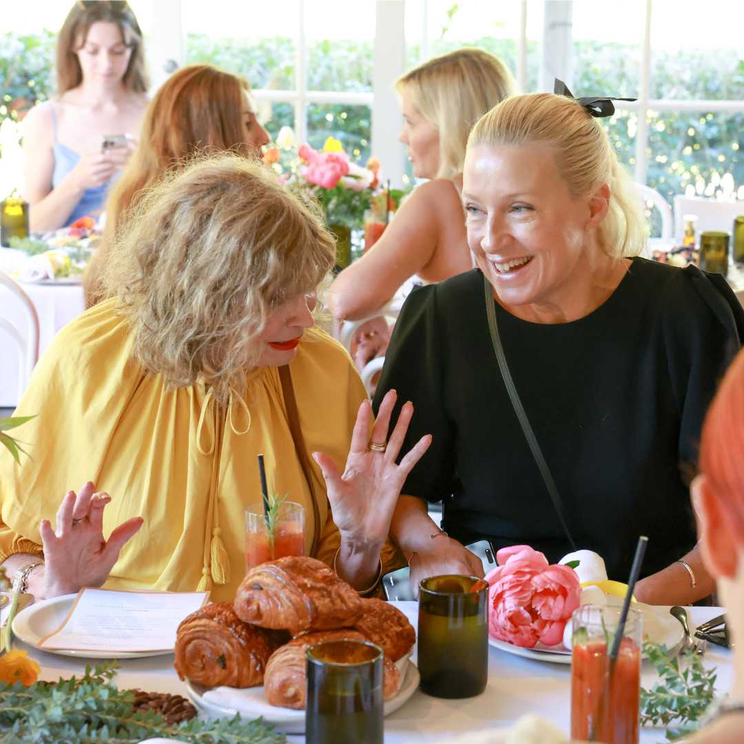 People at a sunny brunch table with pastries, beverages, and floral decorations.