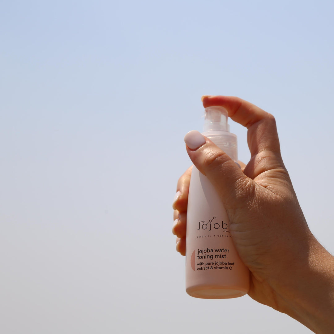 A hand holding a bottle of jojoba water toning mist against a clear sky.