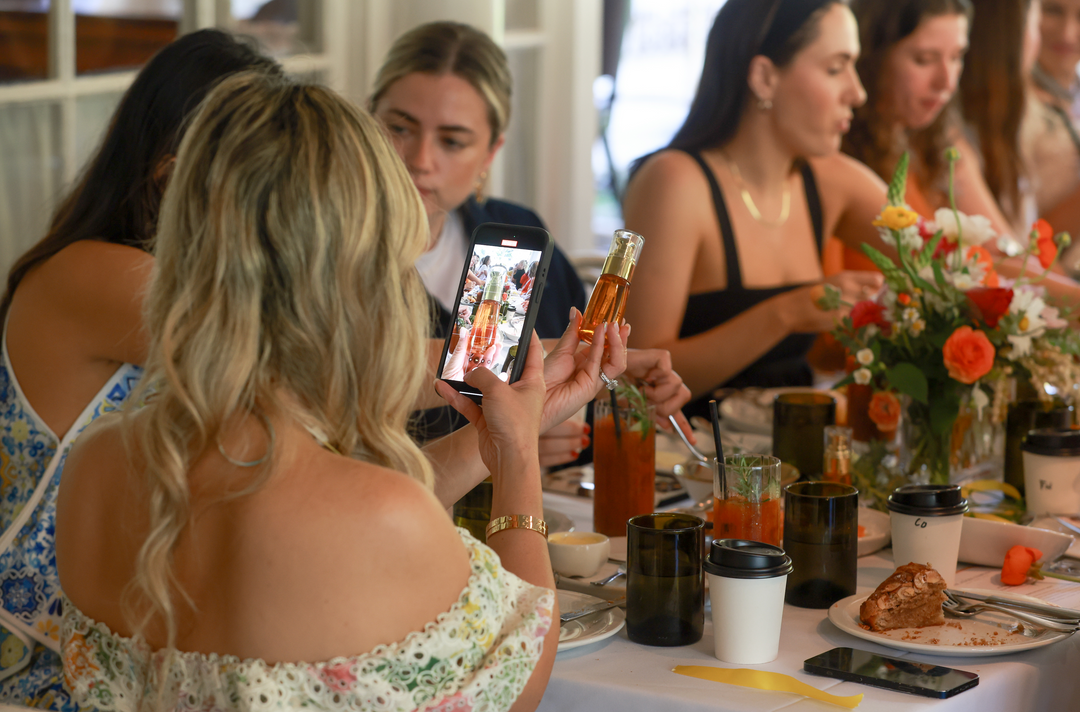 A woman takes a photo of a brunch table with drinks and flowers.