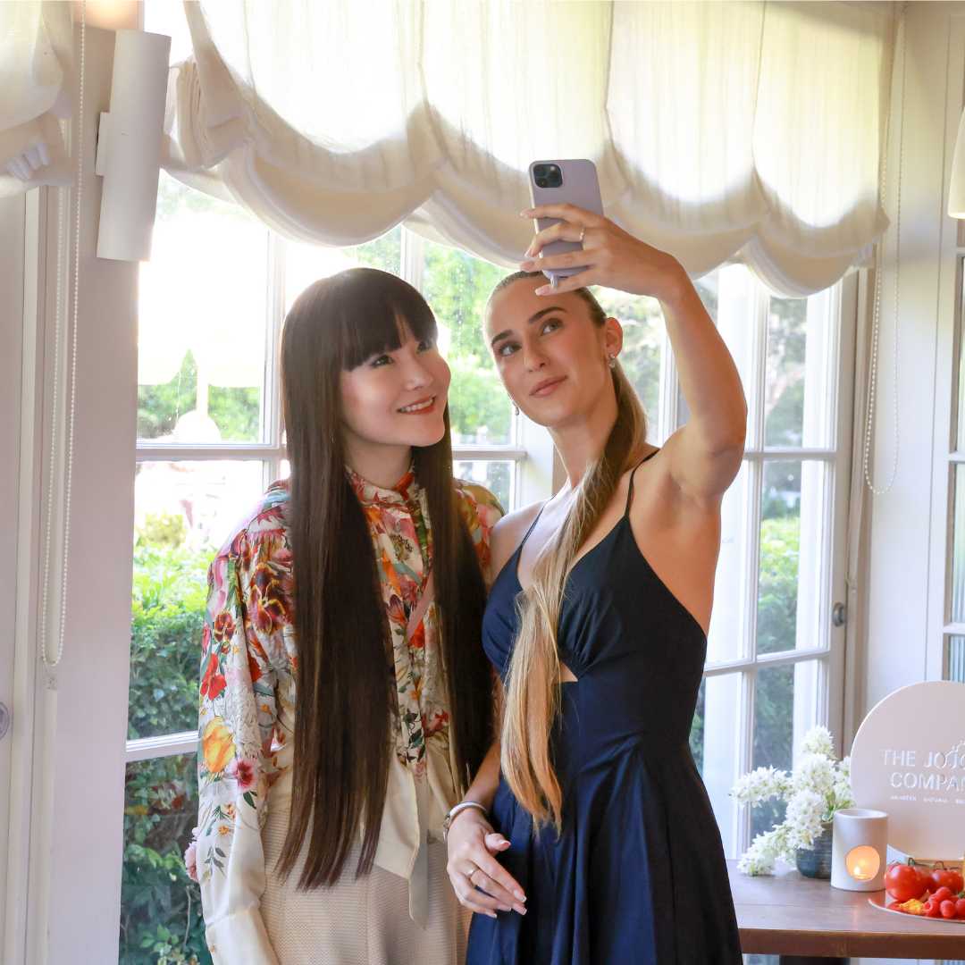 Two individuals taking a selfie in a room with a window, white curtains, and a table with a candle and flowers.